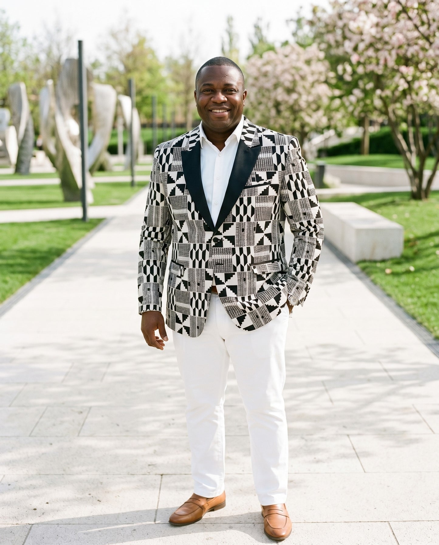 Man wearing a patterned blazer and white pants standing outdoors with trees and sculptures in the background.