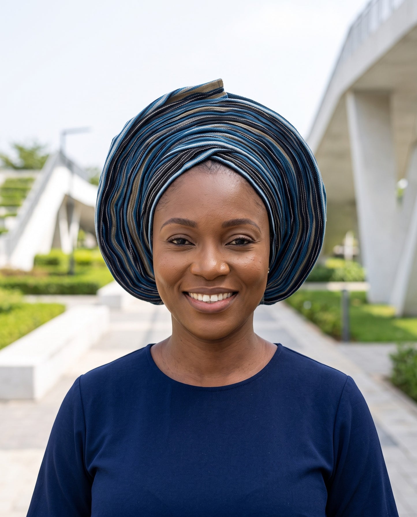 A smiling woman wearing a dark blue and black striped traditional headwrap (dagger) and a navy blue top, standing outdoors in front of a modern white architectural structure.