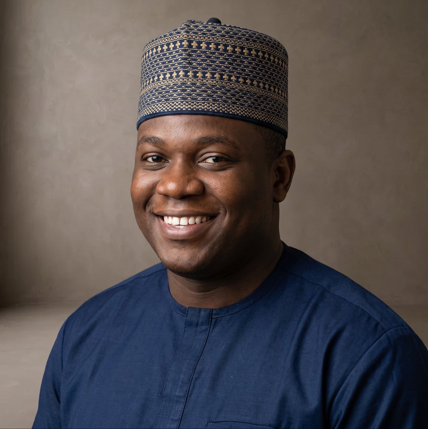 A smiling man wearing a navy blue traditional shirt and a patterned navy and beige cap, photographed against a neutral textured brown studio background.