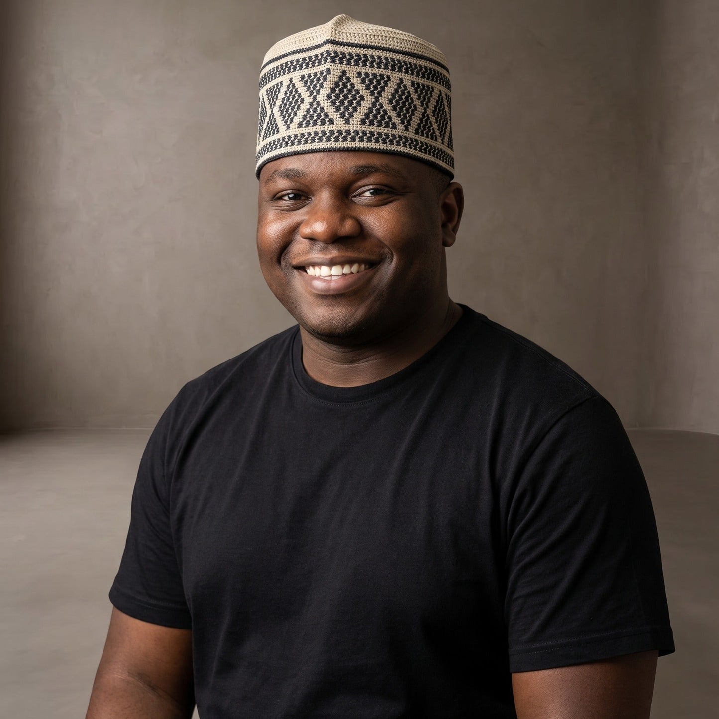 A smiling man wearing a black t-shirt and a cream-colored knit skullcap with a dark geometric pattern, photographed against a neutral textured studio backdrop.