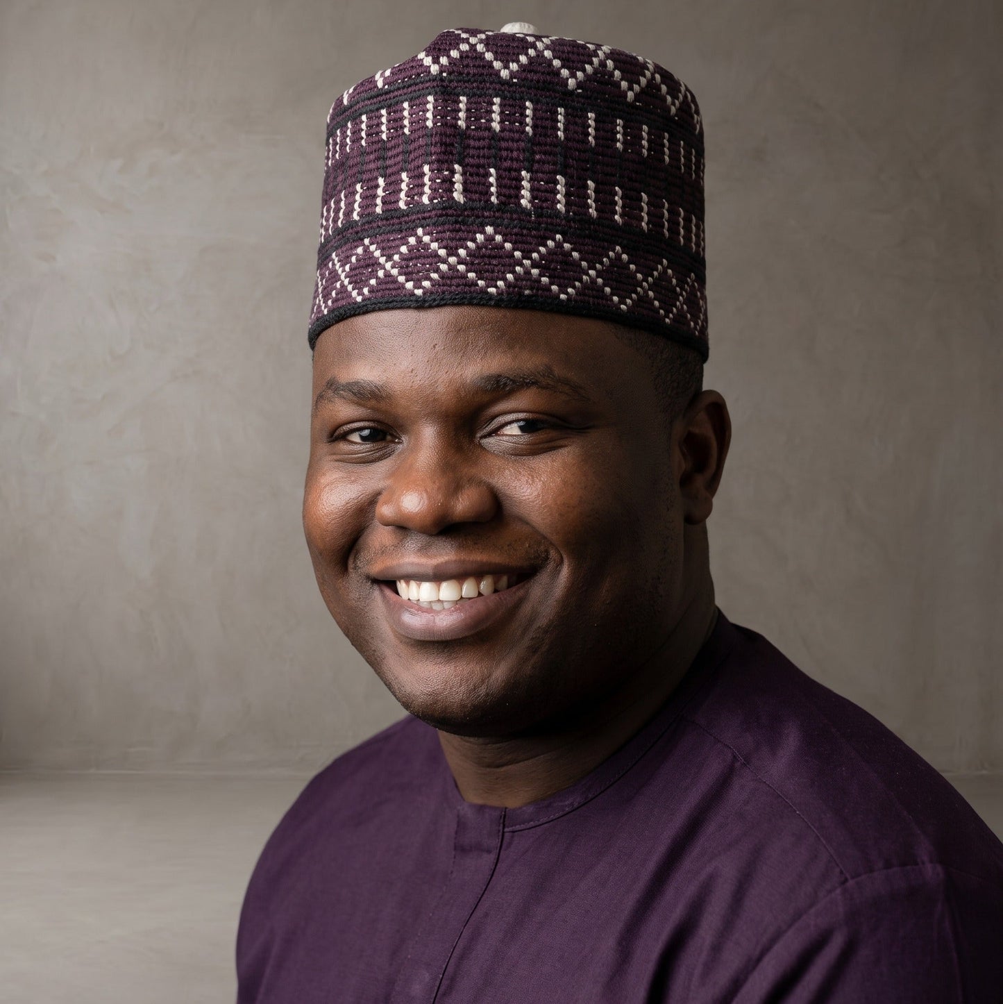 A smiling man wearing a dark purple traditional cap with a white geometric pattern and a matching dark purple shirt, photographed against a neutral textured gray background.