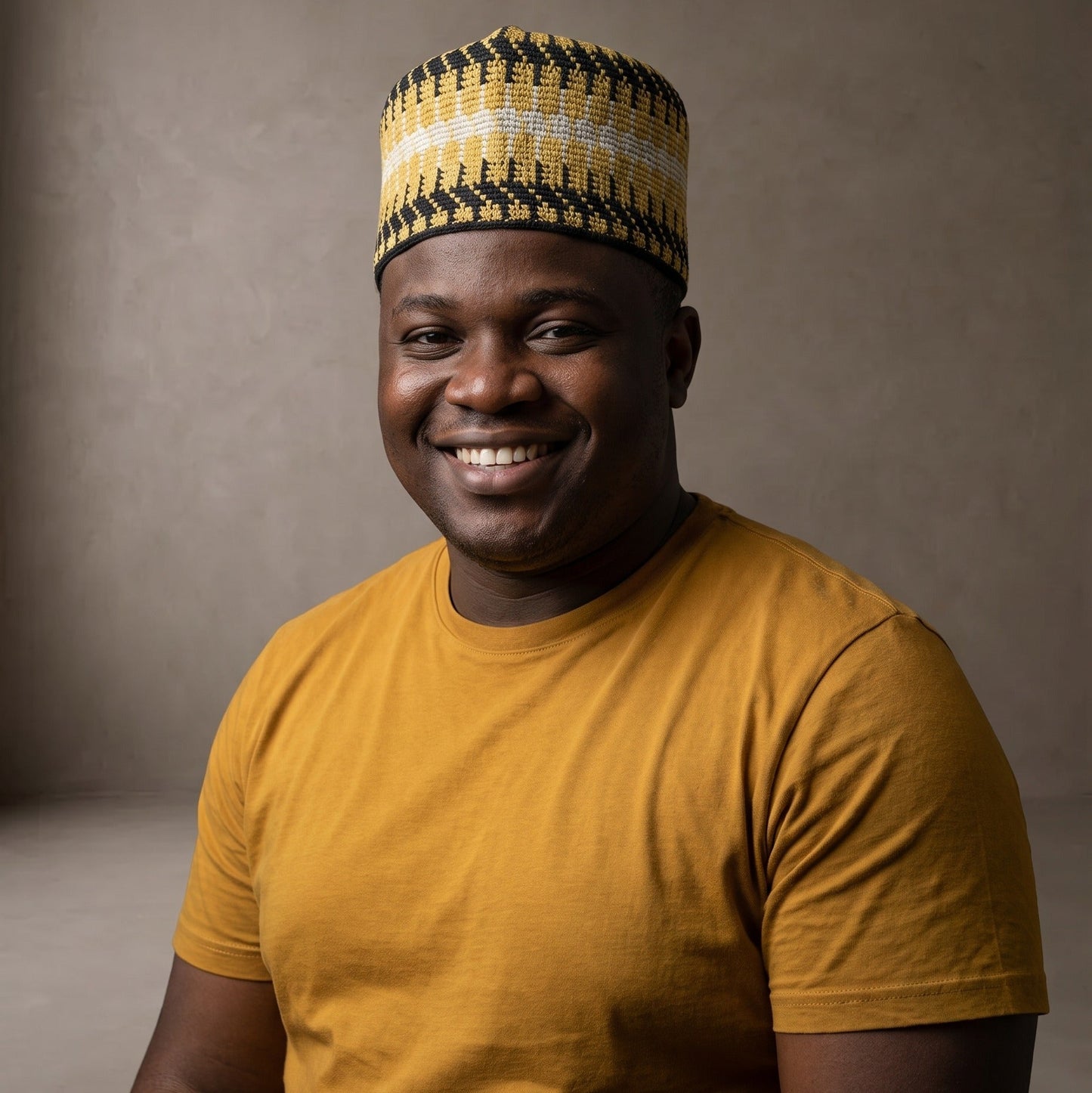 A smiling man wearing a yellow and black patterned knit cap and a mustard yellow t-shirt, photographed against a neutral textured gray studio background.