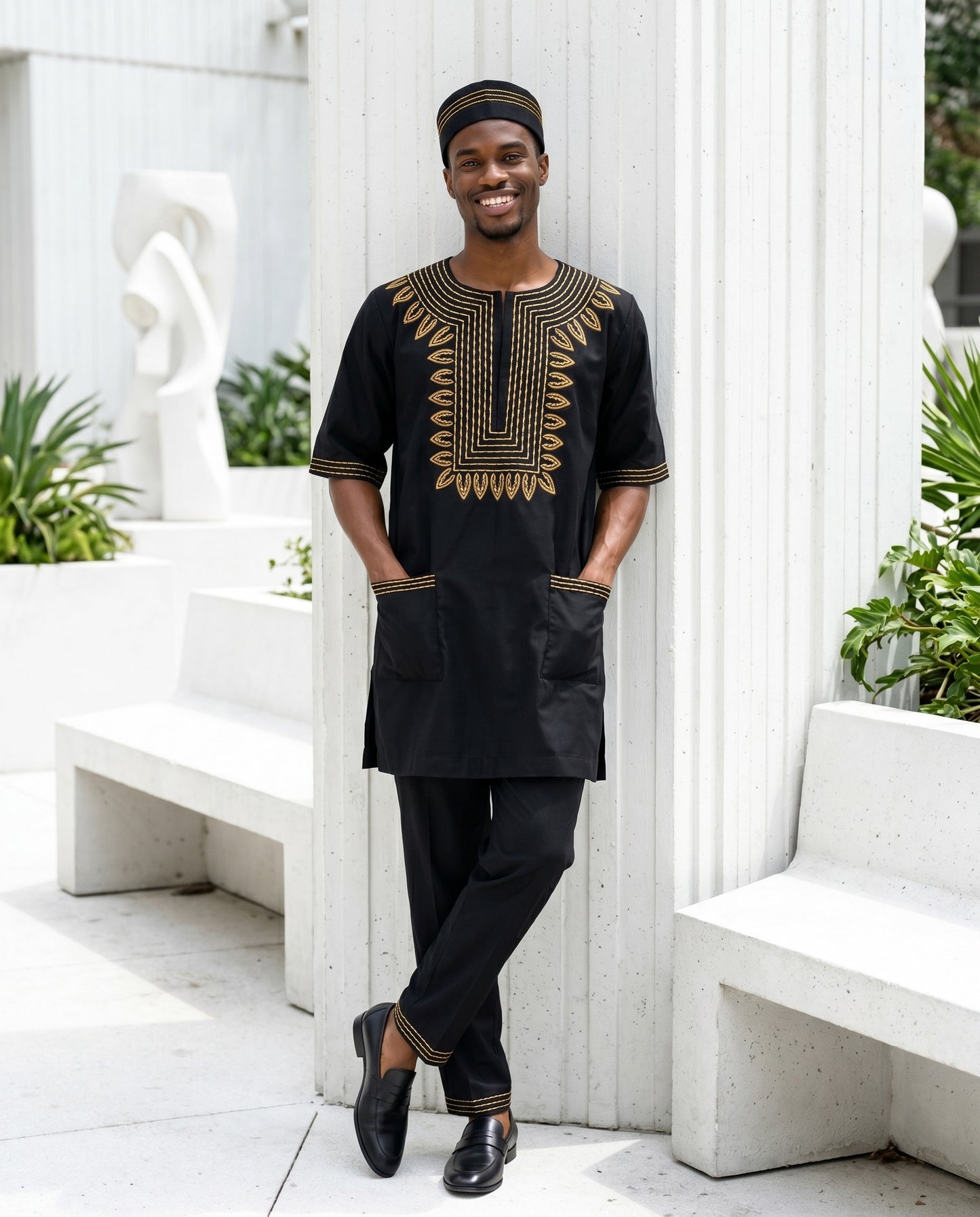Man wearing a black and gold traditional outfit standing in a white outdoor setting.