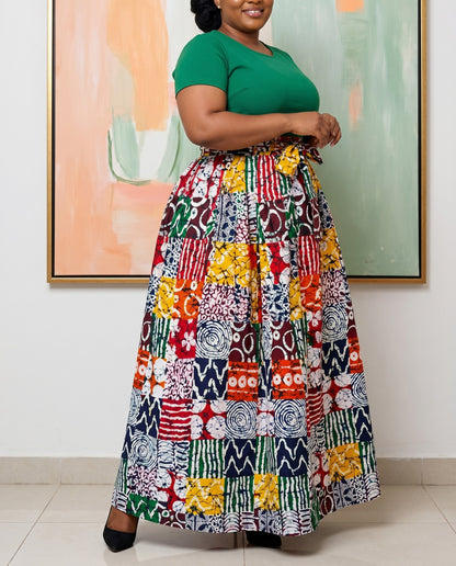 Woman wearing a green top and colorful patterned skirt standing indoors with a abstract painting in the background.