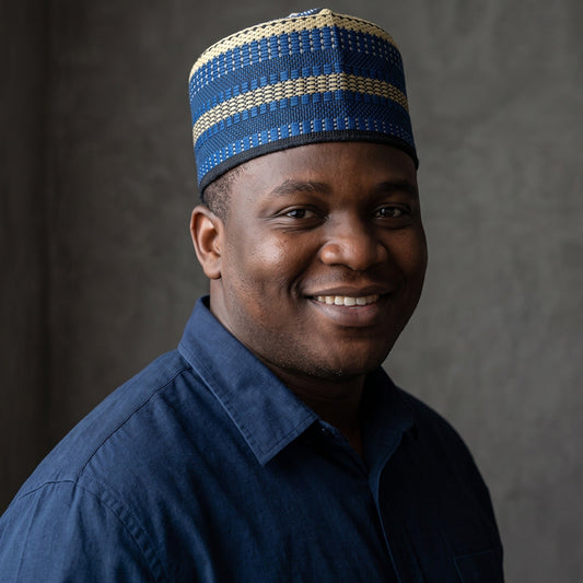 A smiling man wearing a blue and beige patterned knit skullcap and a dark blue button-up shirt, photographed against a textured gray studio background.