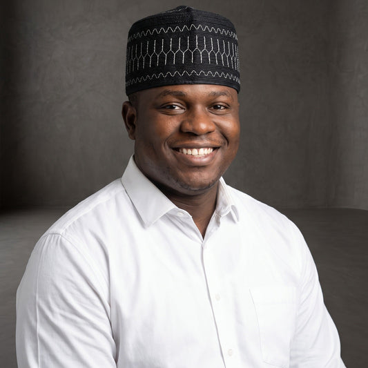A smiling man wearing a white button-up shirt and a black patterned skullcap, photographed against a dark gray studio background.