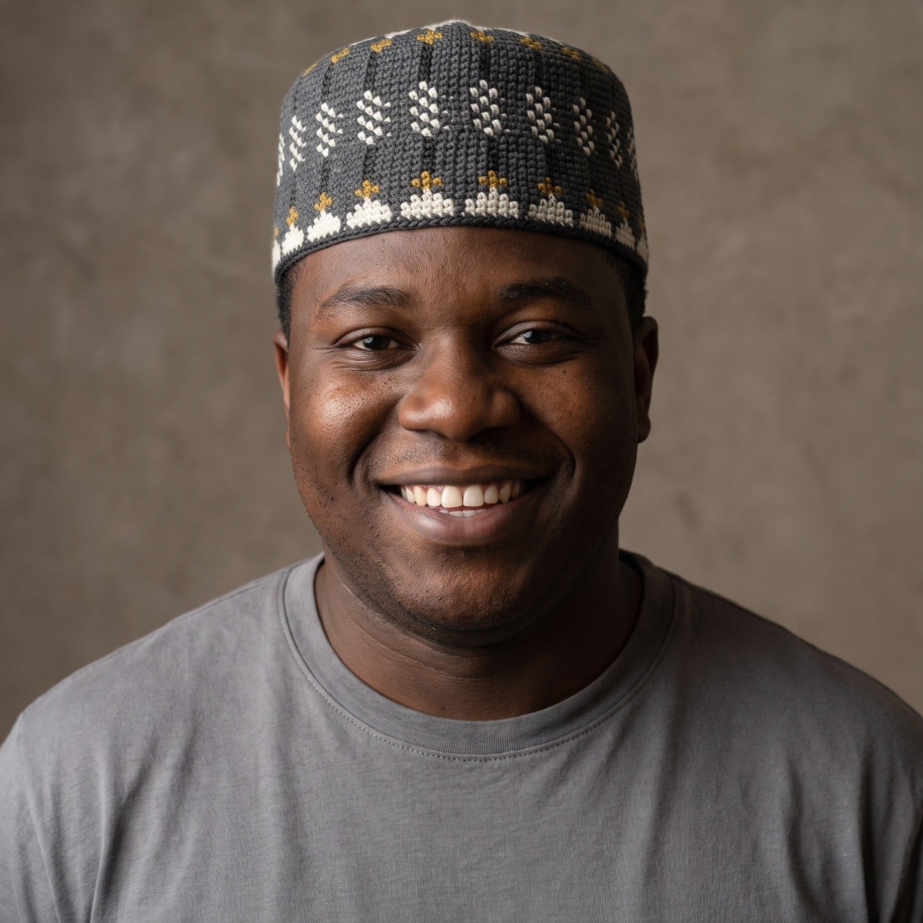 A smiling man wearing a dark gray knit skullcap with white and gold geometric patterns, paired with a gray t-shirt, set against a textured brown studio background.