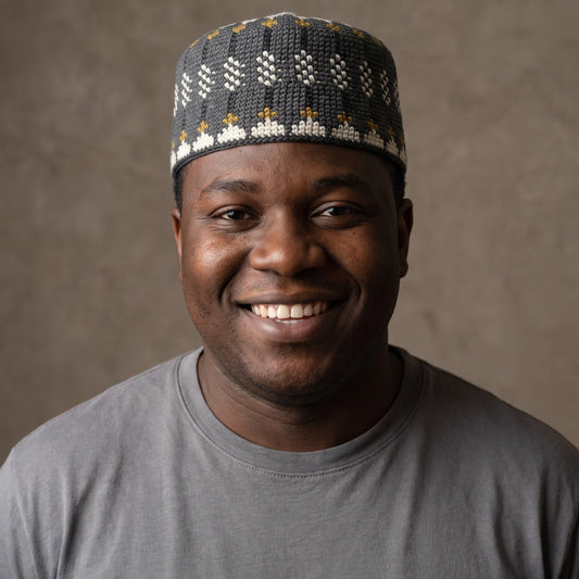 A smiling man wearing a dark gray knit skullcap with white and gold geometric patterns, paired with a gray t-shirt, set against a textured brown studio background.