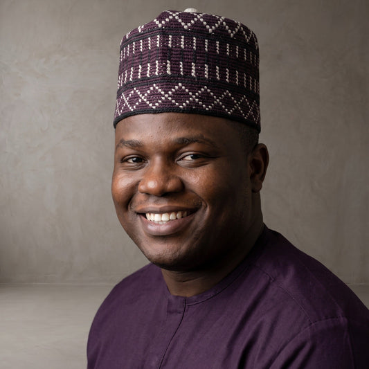 A smiling man wearing a dark purple traditional cap with a white geometric pattern and a matching dark purple shirt, photographed against a neutral textured gray background.