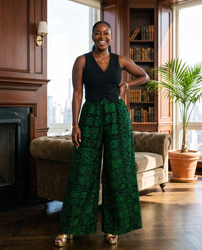 Woman in a black top and green patterned pants standing in a room with bookshelves and a plant.