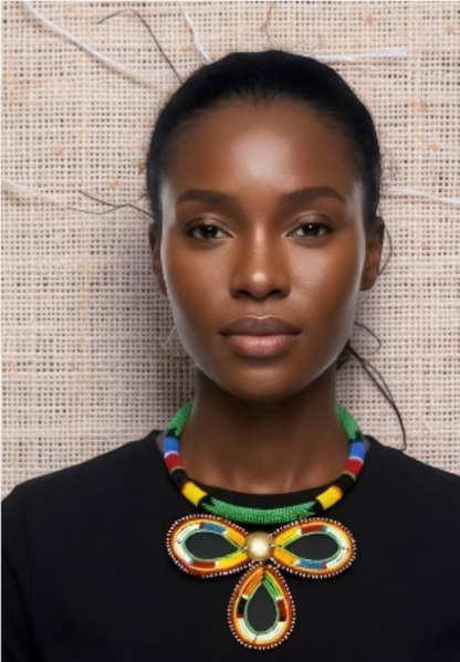 A woman wearing a colorful beaded necklace with a bow-shaped centerpiece and a green beaded collar, paired with a black top against a textured beige background.