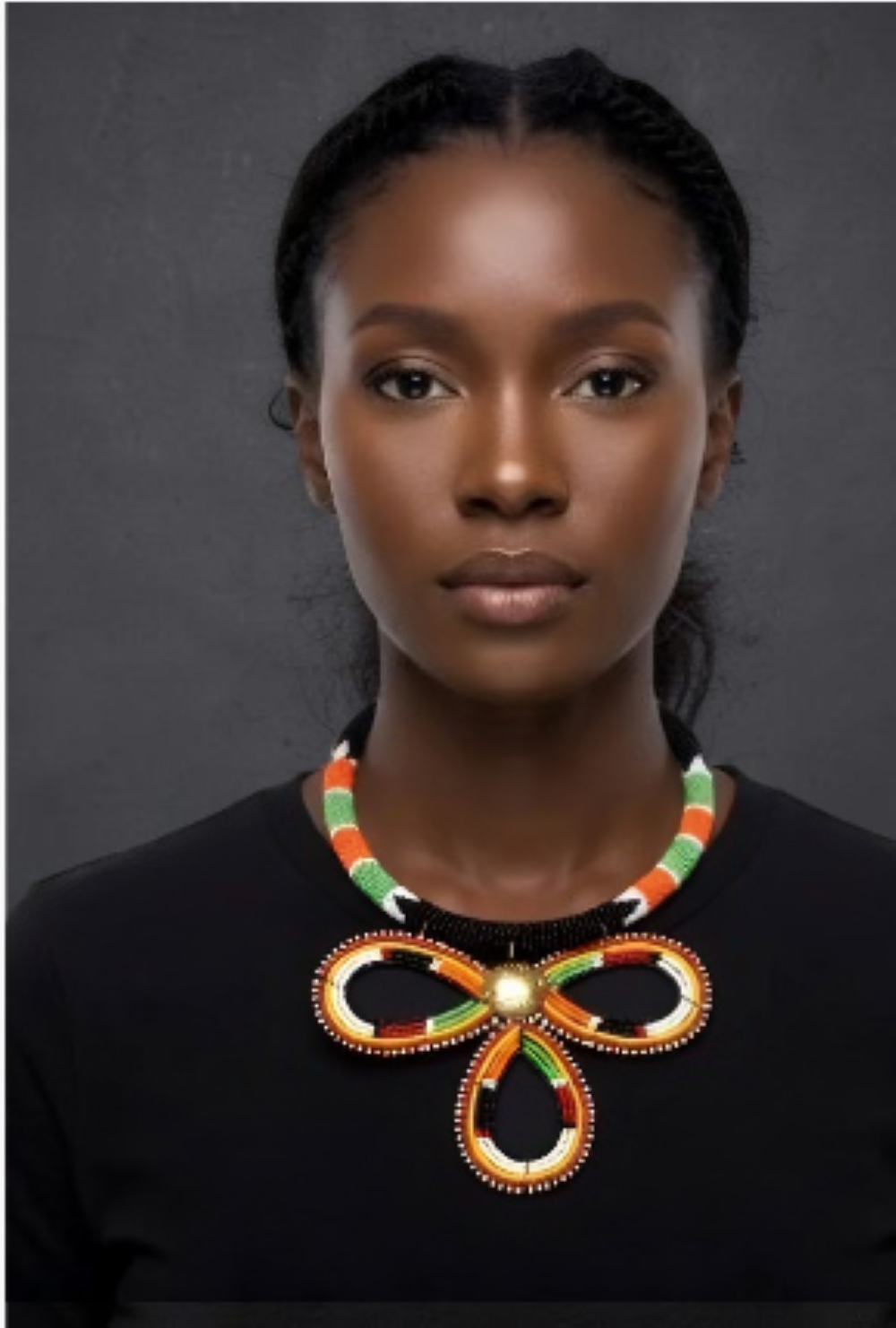 A woman wearing a large, colorful beaded bow-shaped necklace with green, orange, and white beads over a black top, set against a dark gray background.